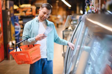 Süpermarketteki dondurulmuş yiyecekleri seçerken elinde alışveriş sepeti tutan ve ambalaj hakkında bilgi okuyan adam