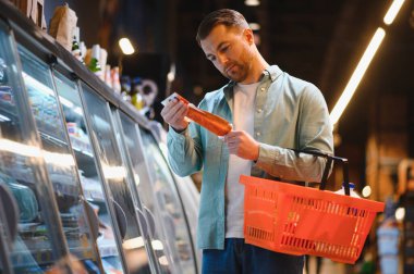 Alışveriş sepeti tutan adam markette soğutulmuş vitrinin yanında paketlenmiş yiyeceklerin etiketini okuyor.