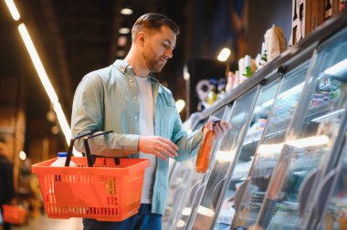 Elinde alışveriş sepeti olan bir adam marketten taze ve dondurulmuş gıda seçenekleriyle dolu bir buzdolabı seçiyor.