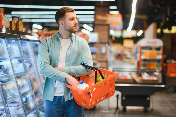 Elinde market poşetleriyle dolu bir sepet tutan bir adam. Parlak bir süpermarket koridorunda duruyor. Etrafı çeşitli ürün ve raflarla çevrili.