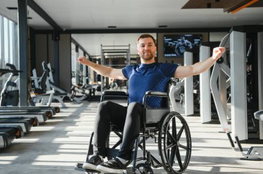 Spor salonunda tekerlekli sandalyede egzersiz yapan engelli bir adam rehabilitasyon sırasında azim ve güç gösterisi yapıyor.