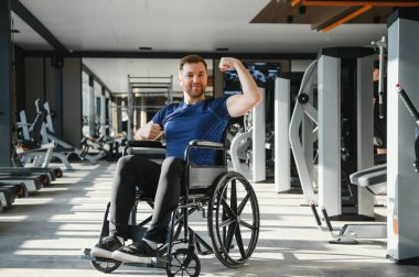 Spor salonunda tekerlekli sandalyede egzersiz yapan, kaslarını gösteren ve gülümseyen güçlü bir sporcu.