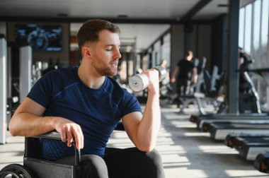Tekerlekli sandalyedeki kararlı erkek sporcu, dambılı kaldırıyor, kolunu parlak ve modern bir spor salonunda çalıştırıyor, rehabilitasyon ve fitness yolculuğuna odaklanıyor.