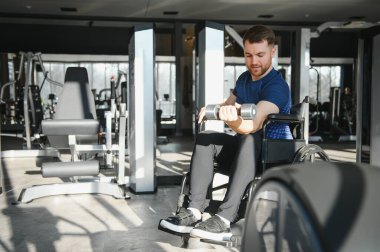 Spor salonunda tekerlekli sandalyede halterle egzersiz yapan engelli bir adam rehabilitasyona ve forma olan bağlılığını gösteriyor.