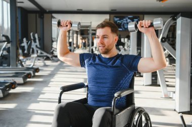 Engelli bir adam modern bir spor salonunda tekerlekli sandalyeyle egzersiz yapıyor, üst vücut gücü ve rehabilitasyonu üzerinde çalışıyor.