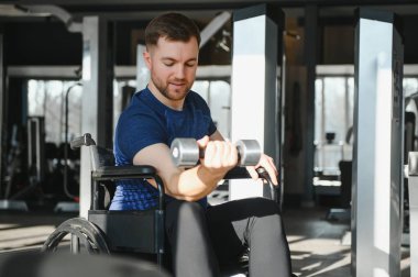 Tekerlekli sandalyedeki adam halterle kol egzersizleri yapıyor, modern bir spor salonunda güç antrenmanı ve rehabilitasyona odaklanıyor.