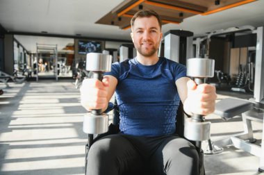 Özürlü gülümseyen sporcu modern bir rehabilitasyon merkezinde tekerlekli sandalyede dambıllarla egzersiz yapıyor.