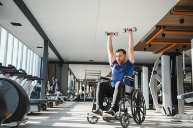 Özürlü bir adam, erkek atlet tekerlekli sandalyede otururken dambıl kaldırıyor, rehabilitasyon ve spor eğitimi için modern spor salonunda egzersiz yapıyor.