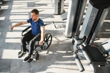 Tekerlekli sandalyedeki adam dambıl kaldırıyor, modern spor salonunda egzersiz yapıyor ve silah eğitiyor, rehabilitasyon ve iyileşme kavramı