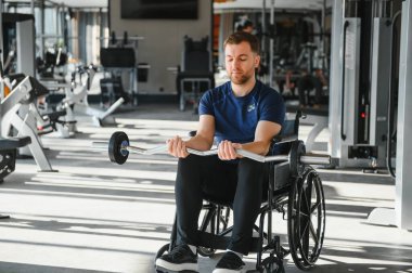 Tekerlekli sandalyede ağırlık kaldıran, modern spor salonunda kollarını çalıştıran, kendini rehabilitasyona ve spor eğitimine adamış bir adam.