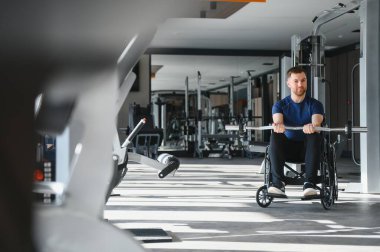 Tekerlekli sandalyedeki erkek sporcu ağırlık kaldırıyor, parlak bir spor salonunda egzersiz yapıyor, rehabilitasyon ve spor eğitimine odaklanıyor.