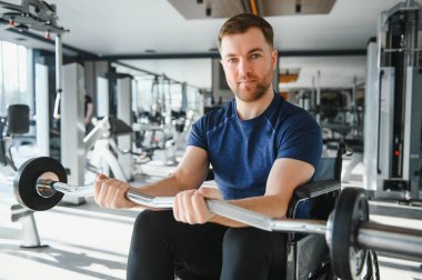 Tekerlekli sandalyedeki adam ağırlık kaldırıyor, spor salonunda kollarını çalıştırıyor, rehabilitasyonu ve iyileşmesi için çalışıyor.