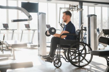 Tekerlekli sandalyedeki erkek sporcu ağırlık kaldırıyor, spor salonunda egzersiz yapıyor, spor ve rehabilitasyona dahil olma gücünü gösteriyor