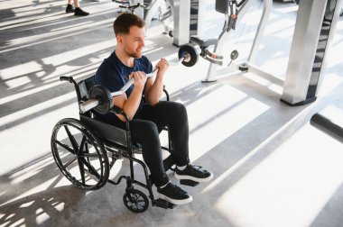 Tekerlekli sandalyede otururken ağırlık kaldıran erkek sporcu, modern spor salonunda rehabilitasyon sırasında egzersiz ve antrenman yapıyor.