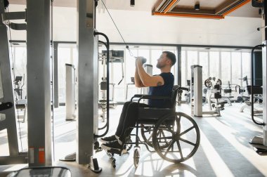 Tekerlekli sandalye kullanan bir adam modern bir spor salonunda ağırlıklarla egzersiz yapıyor. Spor yaparak kapsayıcılık ve rehabilitasyon gösteriyor.
