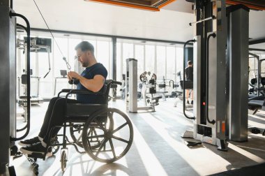 Tekerlekli sandalyedeki bir adam, modern bir spor salonunda dayanıklılık eğitimi alıyor, destekleyici bir çevrede rehabilitasyon ve uygunluğa odaklanıyor.
