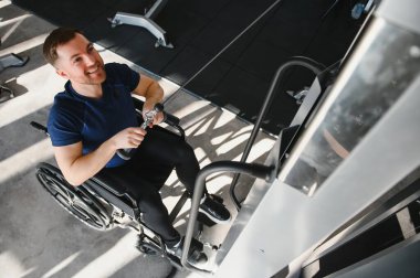 Tekerlekli sandalyedeki gülen adam kablo çekme makinesiyle egzersiz yapıyor, ulaşılabilir spor salonunda rehabilitasyon ve fitness hedeflerine ulaşıyor.
