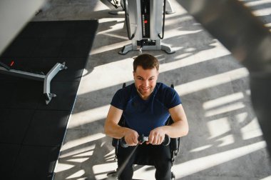 Tekerlekli sandalyede gülümseyen bir adam modern bir spor salonunda kablo ağırlıklarıyla egzersiz yapıyor, rehabilitasyona ve forma bağlılığını gösteriyor.