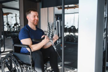 Tekerlekli sandalyedeki gülümseyen adam spor salonunda ağırlıklarla kaslarını çalıştırıyor, spor rehabilitasyonu ve engelliler için adaptasyon eğitimi gösteriyor.