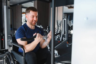 Tekerlekli sandalyede gülümseyen adam modern erişilebilir spor salonunda kablo ağırlıklarıyla egzersiz yapıyor, üst vücut gücü ve rehabilitasyon üzerine çalışıyor.