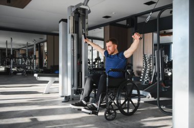 Erkek sporcu adaptasyon ekipmanları kullanıyor, tekerlekli sandalyede ağırlık kaldırıyor, modern bir spor salonunda rehabilitasyon ve spor eğitimi sırasında dayanıklılık ve güç gösteriyor.