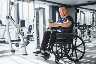 Yeşil bir su şişesinden içen, tekerlekli sandalyede oturan, modern bir spor salonunda egzersiz yapan, gülümseyen bir sporcu.