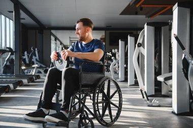 Spor salonunda tekerlekli sandalyede ağırlık egzersizi yapan genç sporcu rehabilitasyon tedavisi görüyor ve spor aktivitelerinden zevk alıyor.