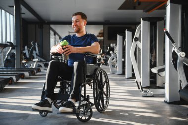 Tekerlekli sandalyede gülümseyen adam spor salonunda egzersiz yaptıktan sonra protein içeceği içiyor, rehabilitasyon ve sağlıklı yaşam tarzı kavramı.