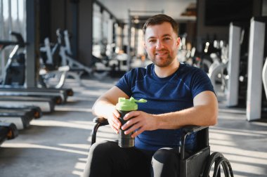 Tekerlekli sandalyedeki mutlu adam protein kokteyli tutuyor, modern rehabilitasyon merkezinde eğitim görüyor.