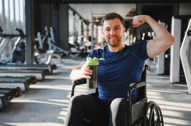 Engelli bir adam elinde protein karıştırıcı tutarak kaslarını esnetiyor, spor salonunda tekerlekli sandalyede oturuyor, gücünü ve kararlılığını gösteriyor.