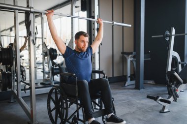 Tekerlekli sandalyede otururken ağırlık kaldıran engelli erkek sporcu spor salonundaki rehabilitasyon egzersizleri sırasında güç ve direnç gösteriyor.