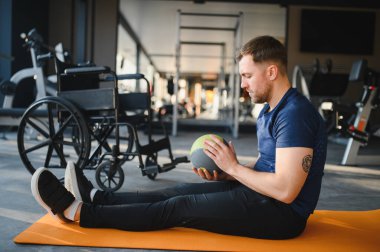 Engelli bir adam spor salonunda rehabilitasyon egzersizleri için ilaç topunu kullanıyor, gücü ve iyileşmeyi teşvik ediyor.