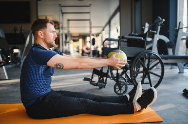 Engelli bir adam jimnastik minderinin üzerinde tıp topu ile egzersiz yapıyor, fitness ve rehabilitasyona bağlılığını gösteriyor.