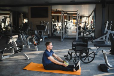 Özürlü bir adam spor salonundaki paspasın üzerinde rehabilitasyon egzersizleri yapıyor, top kullanıyor, tekerlekli sandalyesi yanında, iyileşmeye odaklanıyor.
