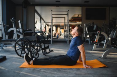 Tekerlekli sandalye kullanıcısı egzersiz yaptıktan sonra spor salonundaki turuncu paspasın üzerinde dinleniyor ve geriniyor.