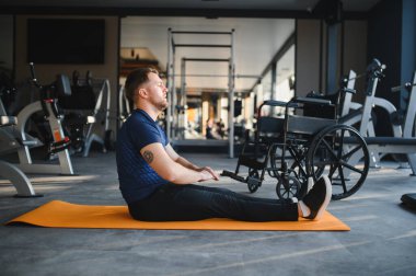 Bir adam paspas üzerinde egzersiz yapıyor, modern bir spor salonunda rehabilitasyona ve iyileşmeye odaklanıyor, tekerlekli sandalyesi yakınlardayken.