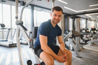 Gülümseyen sporcu modern bir spor salonunda bir eğitim makinesine bağlanıyor. Forma ve sağlıklı bir yaşam tarzına kendini adamış.