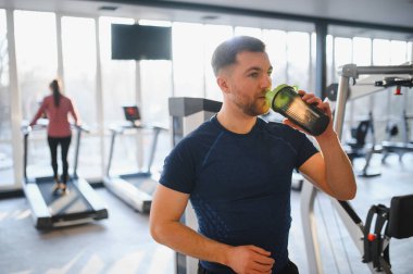 Sakallı bir adam rehabilitasyon merkezinde yoğun bir egzersizden sonra protein içeceği içiyor. İyileşme sürecine ve fitness hedefleri için beslenmeye odaklanıyor.