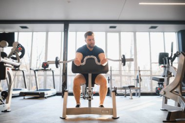 Odaklanmış erkek sporcu, parlak, iyi donanımlı bir spor salonunda, rehabilitasyon egzersizleri sırasında güç ve dayanıklılık göstererek, bir ez çubuğuyla biseps yapıyor.