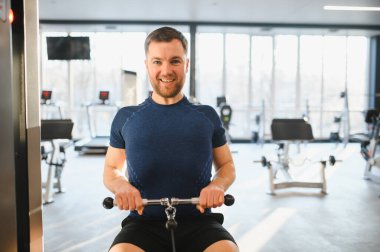 Gülümseyen bir adam rehabilitasyon sırasında oturmuş bir kablo sıra makinesiyle meşgul oluyor. Spor salonundaki iyileşme ve güce odaklanıyor.