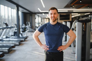 Gülümseyen fitness hocası modern bir spor salonunda elleri kalçasında, fitness eğitiminde kendine güvenini ve uzmanlığını gösteriyor.