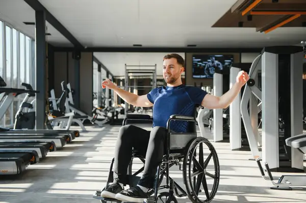 Tekerlekli sandalyedeki genç adam, parlak, modern bir spor salonunda rehabilitasyon için vücut egzersizleri yapıyor. Kararlılık ve güç gösterisi yapıyor.