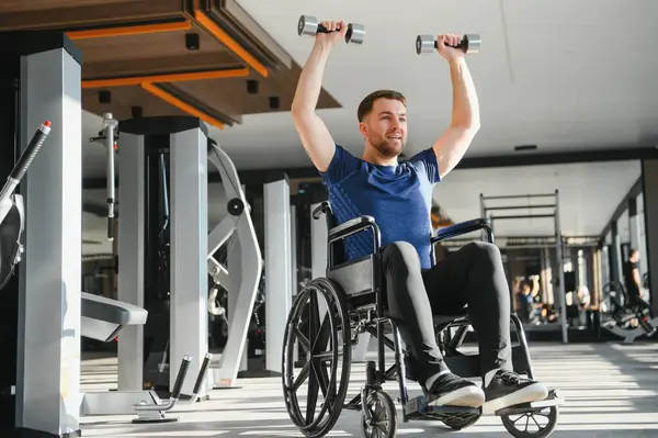 Tekerlekli sandalyedeki gülümseyen adam rehabilitasyon egzersizleri sırasında ağırlık kaldırıyor, ulaşılabilir bir spor salonunda azim ve güç gösteriyor.