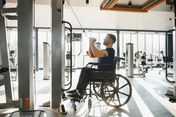 Tekerlekli sandalye kullanan bir adam modern bir spor salonunda ağırlıklarla egzersiz yapıyor. Spor yaparak kapsayıcılık ve rehabilitasyon gösteriyor.