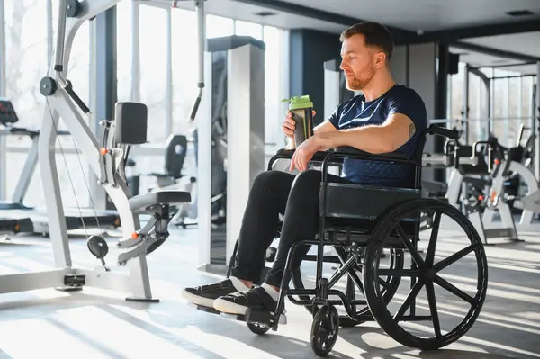 Tekerlekli sandalyedeki beyaz adam su şişesinden içiyor, modern, parlak bir spor salonunda egzersiz yaptıktan sonra dinleniyor, rehabilitasyonu, sporu destekliyor ve zorlukların üstesinden geliyor.