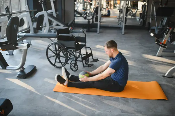 Spor salonundaki turuncu paspasın üzerinde antrenman yaparken sağlık topu kullanan, tekerlekli sandalyeli, rehabilitasyonu destekleyen ve spor salonunu kapsayan