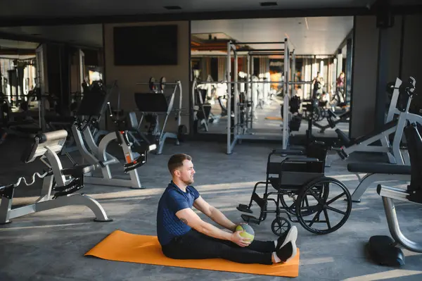 Özürlü bir adam spor salonundaki paspasın üzerinde rehabilitasyon egzersizleri yapıyor, top kullanıyor, tekerlekli sandalyesi yanında, iyileşmeye odaklanıyor.
