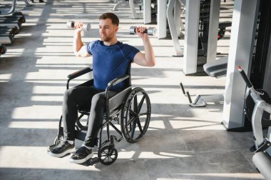 Engelli bir adam tekerlekli sandalyede dambıllarla egzersiz yapıyor. Modern bir spor salonunda rehabilitasyon sırasında azim ve güç gösteriyor.