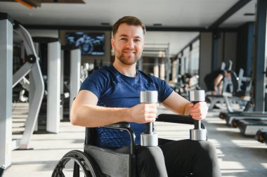 Tekerlekli sandalyeli bir adam modern bir spor salonunda dambıllarla egzersiz yapıyor spor rehabilitasyonunu ve engelli bireyler için kapsayıcı uygunluğu vurguluyor.