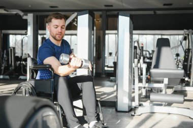 Tekerlekli sandalyesinde otururken ağırlık kaldıran engelli bir sporcu rehabilitasyon ve fitness rutininin bir parçası olarak modern bir spor salonunda egzersiz yapıyor.